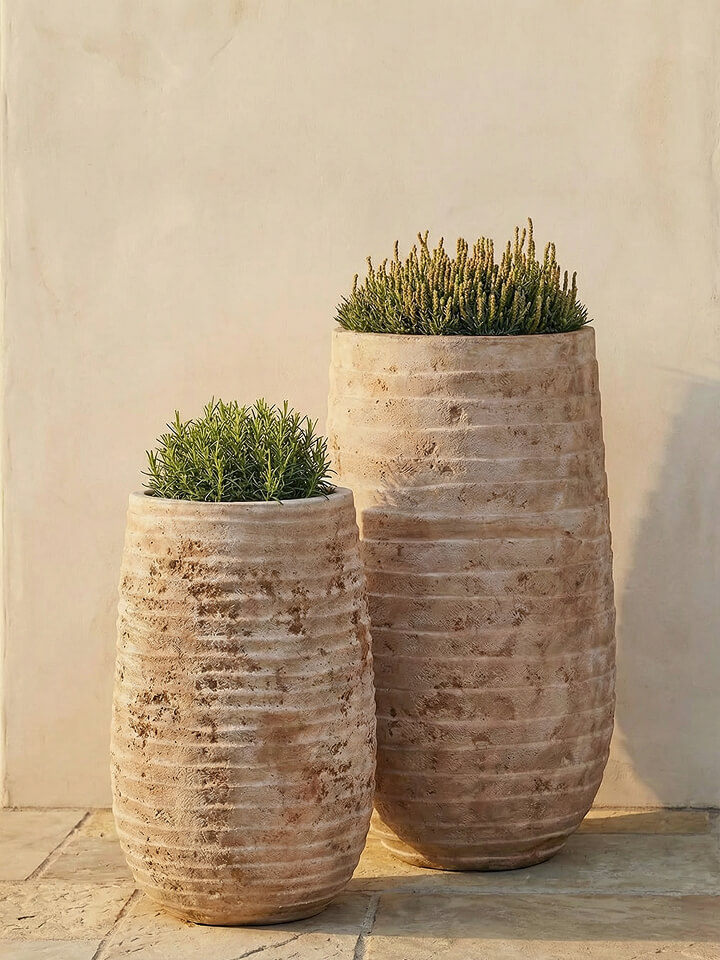 Dos jardineras de piedra texturizada de diferentes alturas contienen cada una una pequeña planta verde, colocadas contra una pared lisa de color beige sobre un suelo de baldosas.