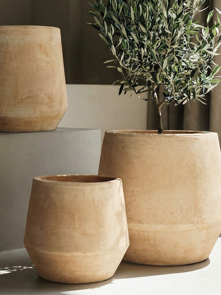 Se exponen tres grandes macetas de cerámica de color marrón claro, una de las cuales contiene una frondosa planta verde. Las macetas tienen un acabado liso y mate y están colocadas sobre un fondo neutro.