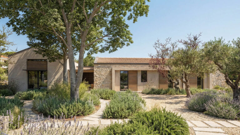Casa de una sola planta con exterior de piedra y estuco, contraventanas de madera y tejado de tejas, rodeada de árboles, arbustos y un camino de piedra en un jardín paisajista.