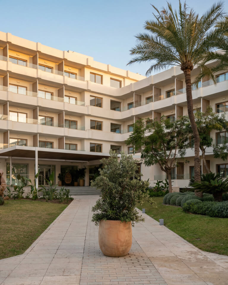 Un moderno hotel de cuatro plantas con balcones, palmeras y zonas verdes ajardinadas, visto desde una pasarela de piedra que conduce a la entrada.