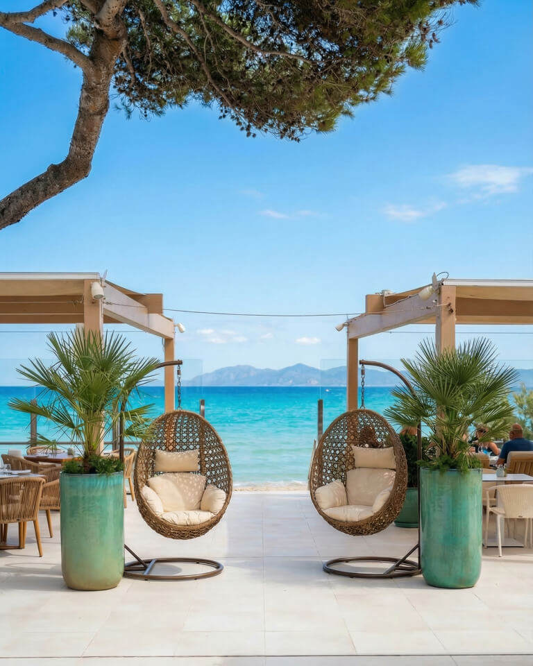 Dos sillas colgantes de mimbre con cojines están colocadas en un patio frente al océano, flanqueado por palmeras en macetas. Cerca, los asientos al aire libre ofrecen unas vistas perfectas del cielo azul despejado, ideales para relajarse o disfrutar de los restaurantes locales.