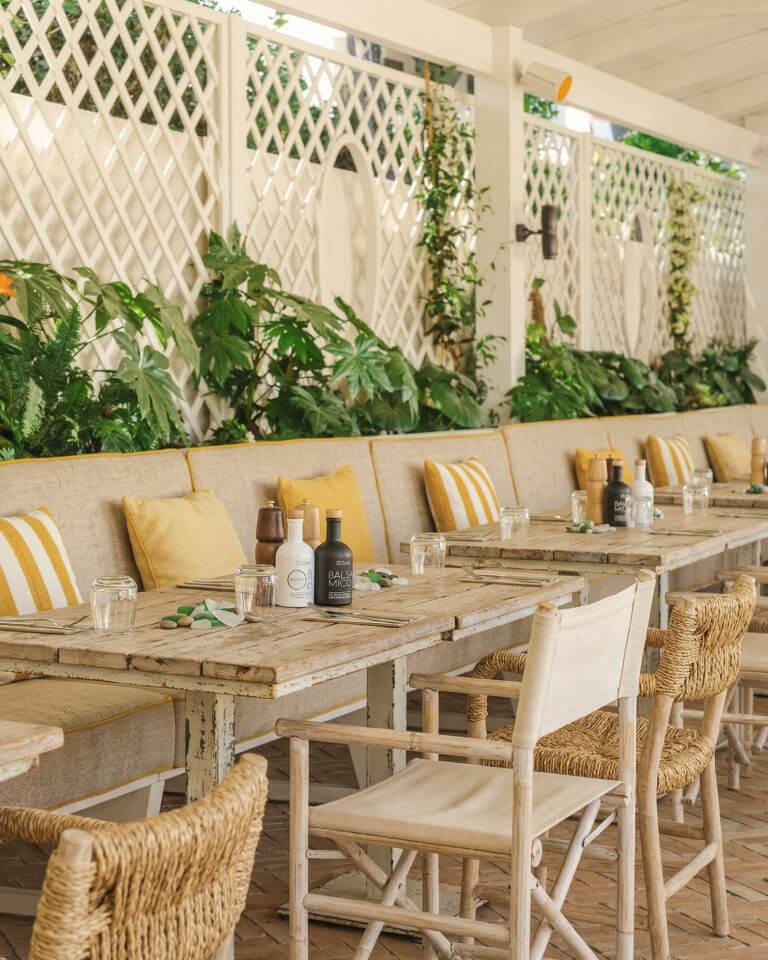 Restaurante al aire libre con mesas de madera, bancos acolchados con cojines amarillos y blancos, sillas tejidas y vegetación decorativa a lo largo de una pared de celosía blanca: un espacio acogedor que suele encontrarse en los restaurantes con estilo.