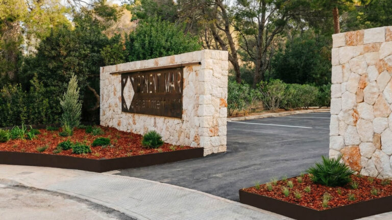 Entrada a un aparcamiento con un muro de piedra y un cartel metálico, rodeado de plantas y árboles al fondo.