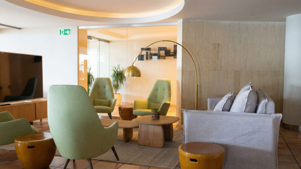 Moderna zona de estar con sillones verdes y grises, un sofá, mesas de madera y plantas de interior, con una lámpara de pie y decoración neutra.