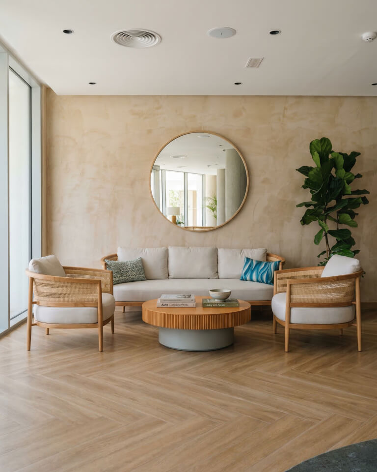 Moderno salón en residencias privadas con sofá beige, dos sillones, mesa de centro redonda de madera, gran espejo de pared redondo, planta en maceta y decoración neutra.