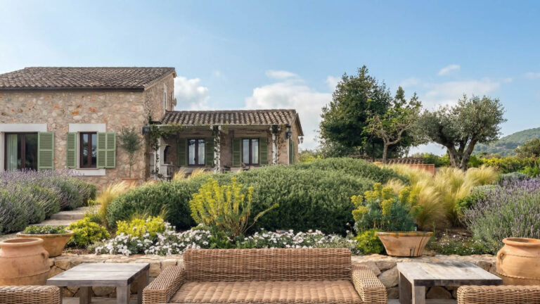 Casa de piedra con tejado de tejas y contraventanas verdes, rodeada de exuberantes jardines, asientos de mimbre al aire libre y plantas en macetas bajo un cielo azul despejado.
