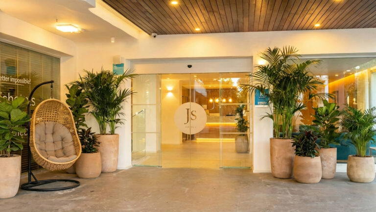 Moderna entrada de hotel con grandes puertas de cristal, plantas en macetas, una silla colgante y el logotipo "JS" expuesto en las puertas de cristal. En el interior se aprecia una cálida iluminación.