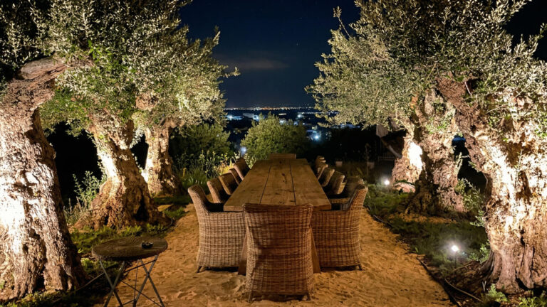 Una larga mesa de comedor de madera con sillas de mimbre está colocada al aire libre sobre suelo arenoso entre olivos iluminados por la noche, con vistas a las luces de la ciudad en la distancia.
