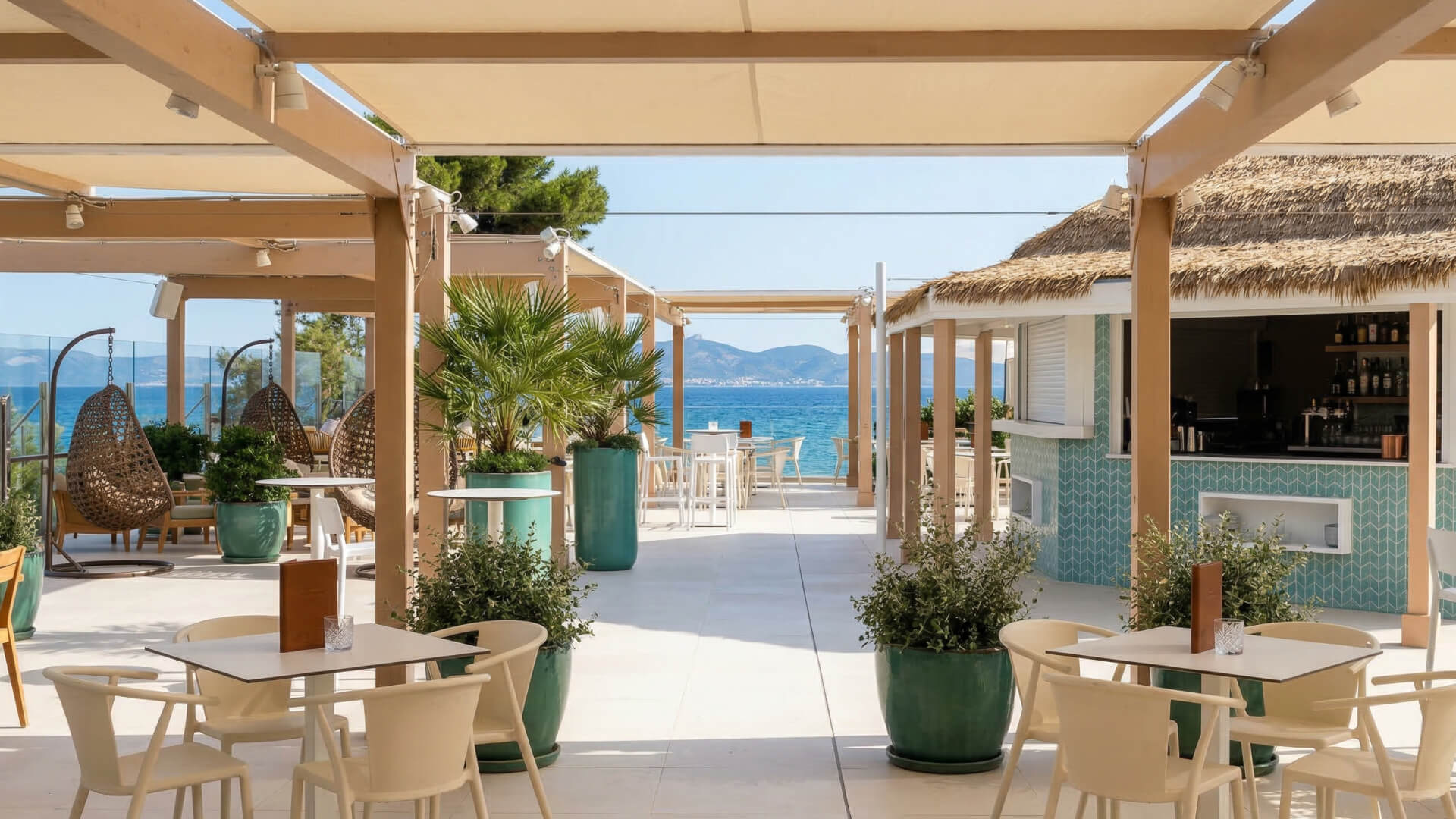 Terraza de restaurante al aire libre con decoración moderna, plantas en macetas, sillas de mimbre y una impresionante vista del mar y las montañas lejanas: uno de los restaurantes más acogedores para disfrutar de una cena bajo un cielo despejado.