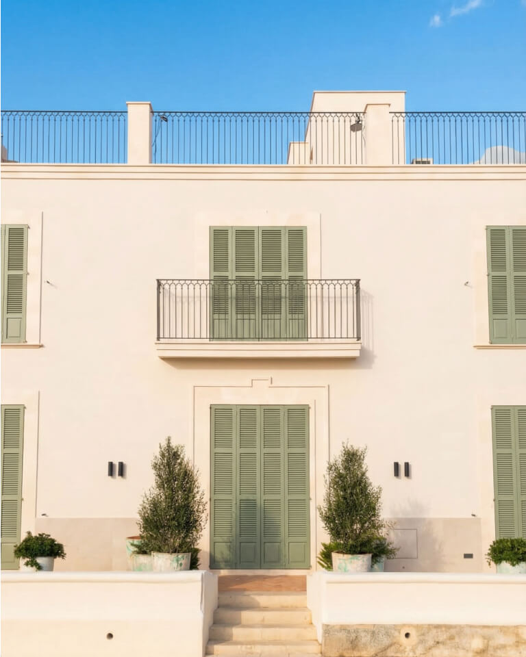 Un edificio de residencias privadas de color crema con contraventanas verdes y un pequeño balcón; dos plantas en macetas flanquean la entrada, bajo un cielo azul despejado.